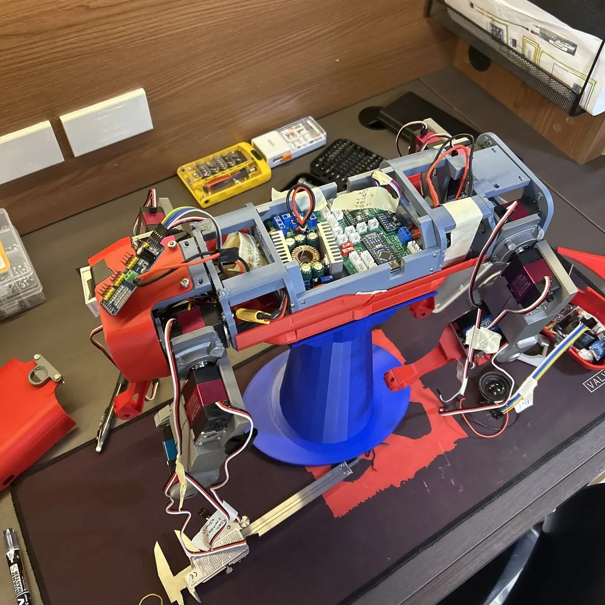 The same quadruped on a workbench with its top shell removed, held up on a blue printed stand. Its grey internal frame carries several circuit boards and a loom of wiring, a servo sits at each leg joint, and a pair of calipers and a screwdriver bit set lie on the mat beside it.