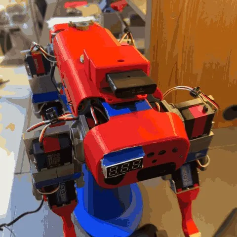 A red, blue and white 3D-printed quadruped robot standing on a desk, filmed from a camera circling it. Servos sit at each leg joint, a small numeric display faces out of the red body, and wiring loops between the panels.