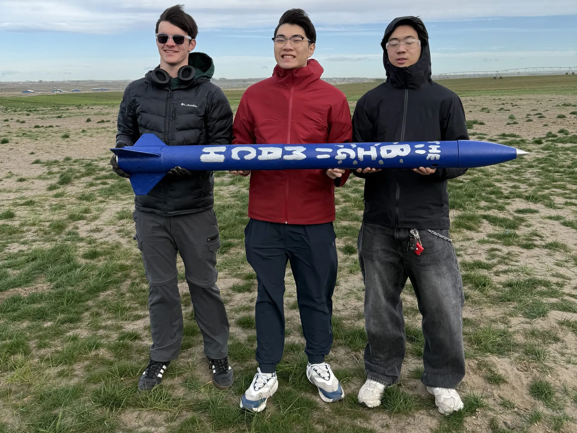 Three team members standing shoulder to shoulder in a flat grass field under a broad, overcast sky, together holding a blue rocket horizontally at waist height. White block lettering runs the length of its body.